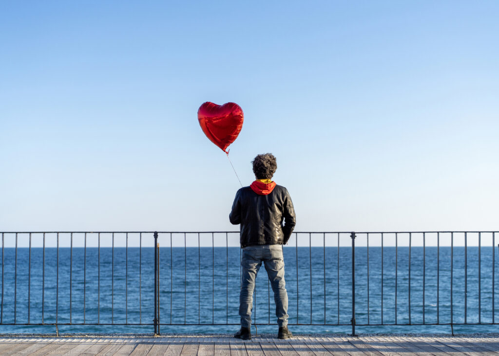 Einsamer Mann, der einen roten herzförmigen Ballon am Strand hält Männerherzen Männer dialoge 11-2025